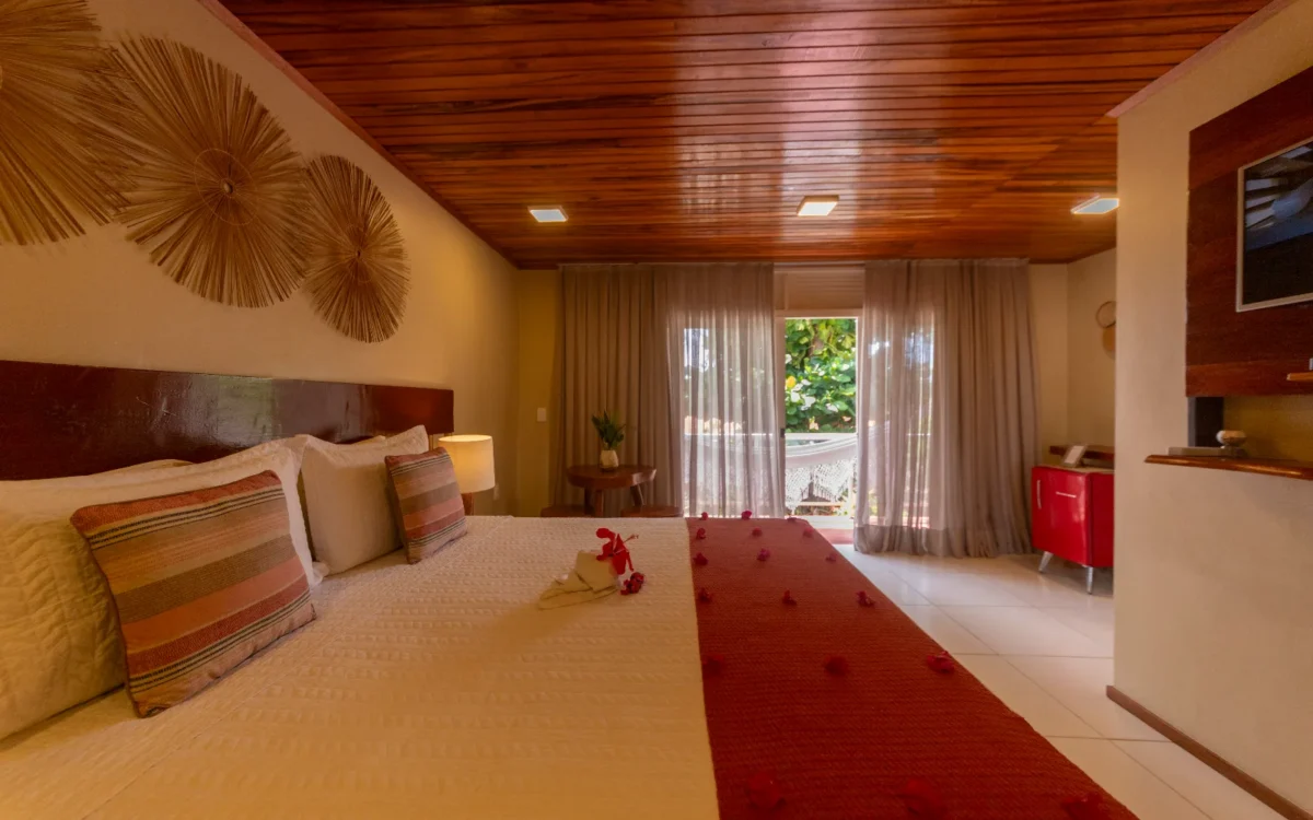 Quarto de hotel com cama de casal, cabeceira vermelha, teto de madeira, cortinas bege, flores vermelhas e porta aberta para varanda com vista para vegetação