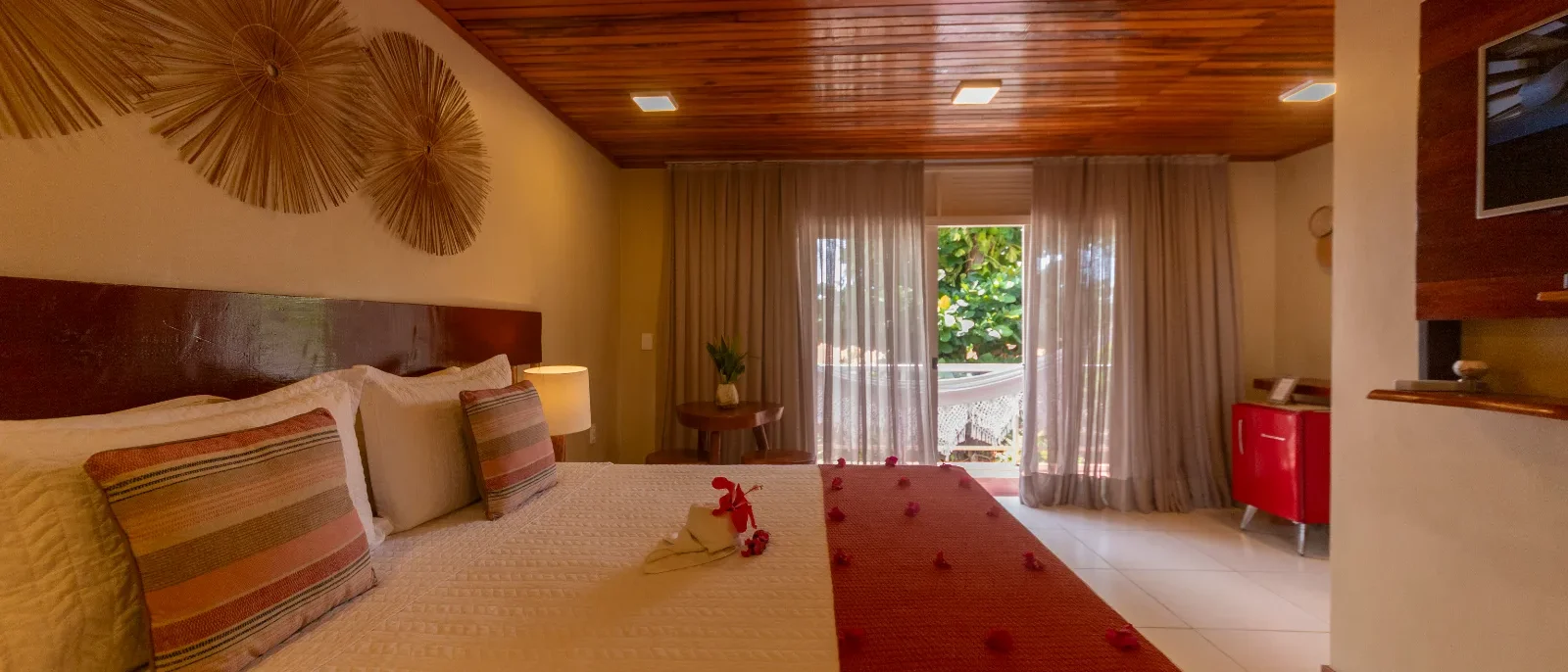 Quarto de hotel com cama de casal, cabeceira vermelha, teto de madeira, cortinas bege, flores vermelhas e porta aberta para varanda com vista para vegetação