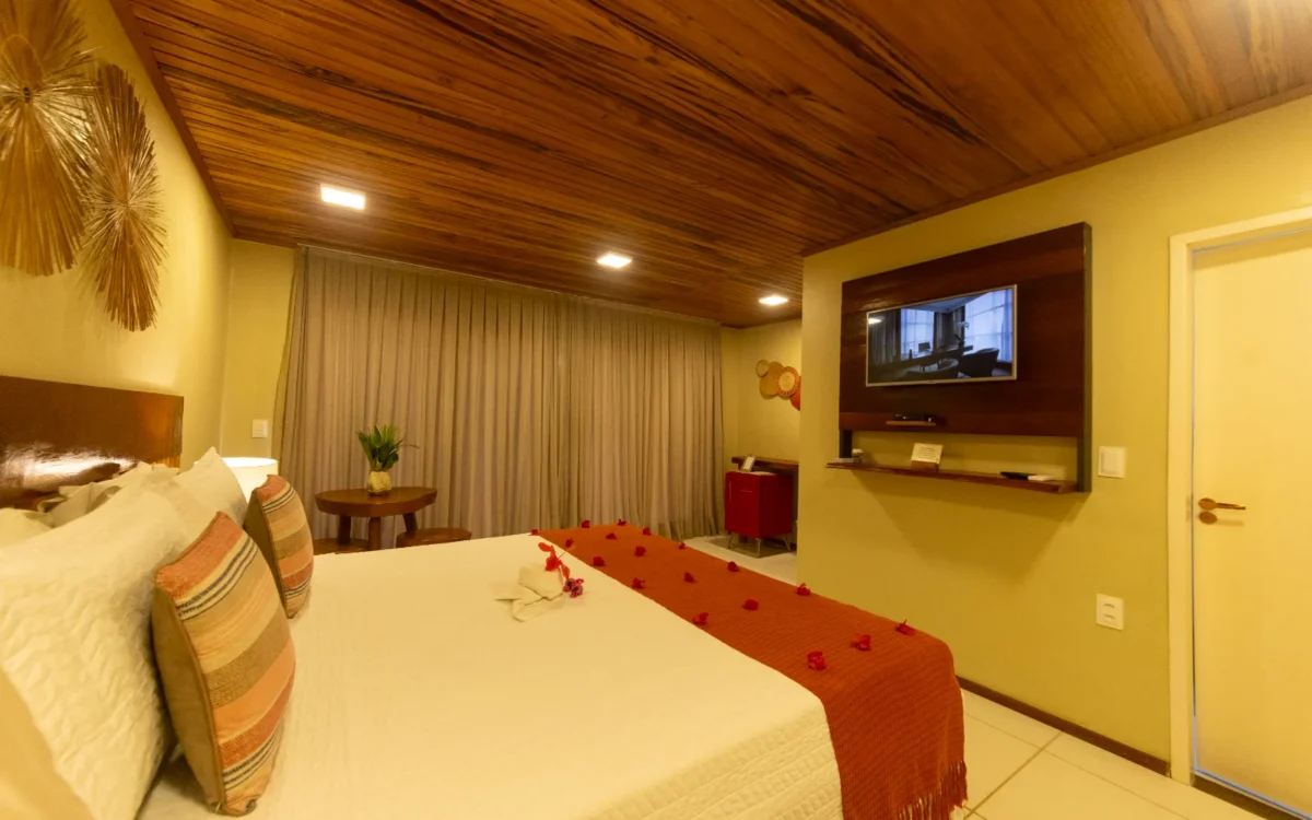 Quarto de hotel com cama de casal, cabeceira em madeira, parede amarela, teto em madeira, TV na parede, cortinas bege e decoração em tons quentes