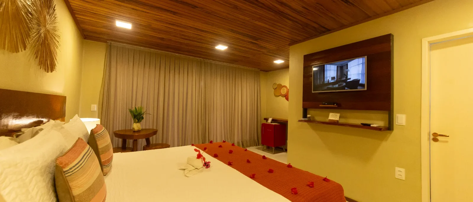 Quarto de hotel com cama de casal, cabeceira em madeira, parede amarela, teto em madeira, TV na parede, cortinas bege e decoração em tons quentes