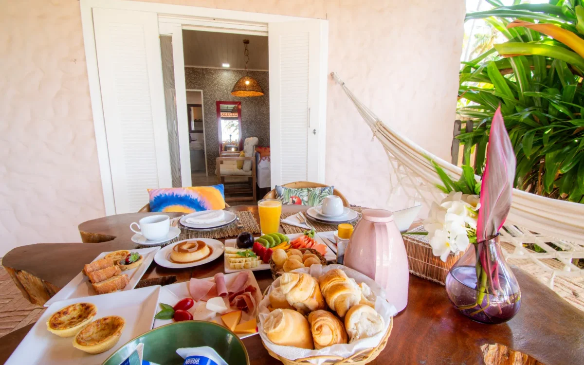 Mesa com café da manhã contendo pães, croissants, frutas, presunto, sucos, café e flores, com porta aberta ao fundo mostrando interior da casa