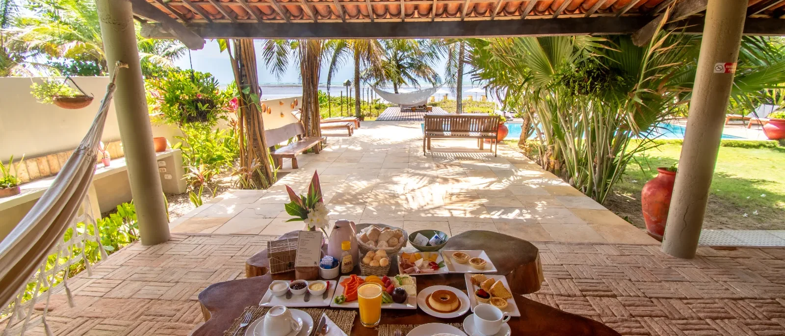 Mesa com café da manhã servido sob pérgolado com telhas de terracota, vista para jardim com palmeiras, piscina e estrutura de lazer ao fundo