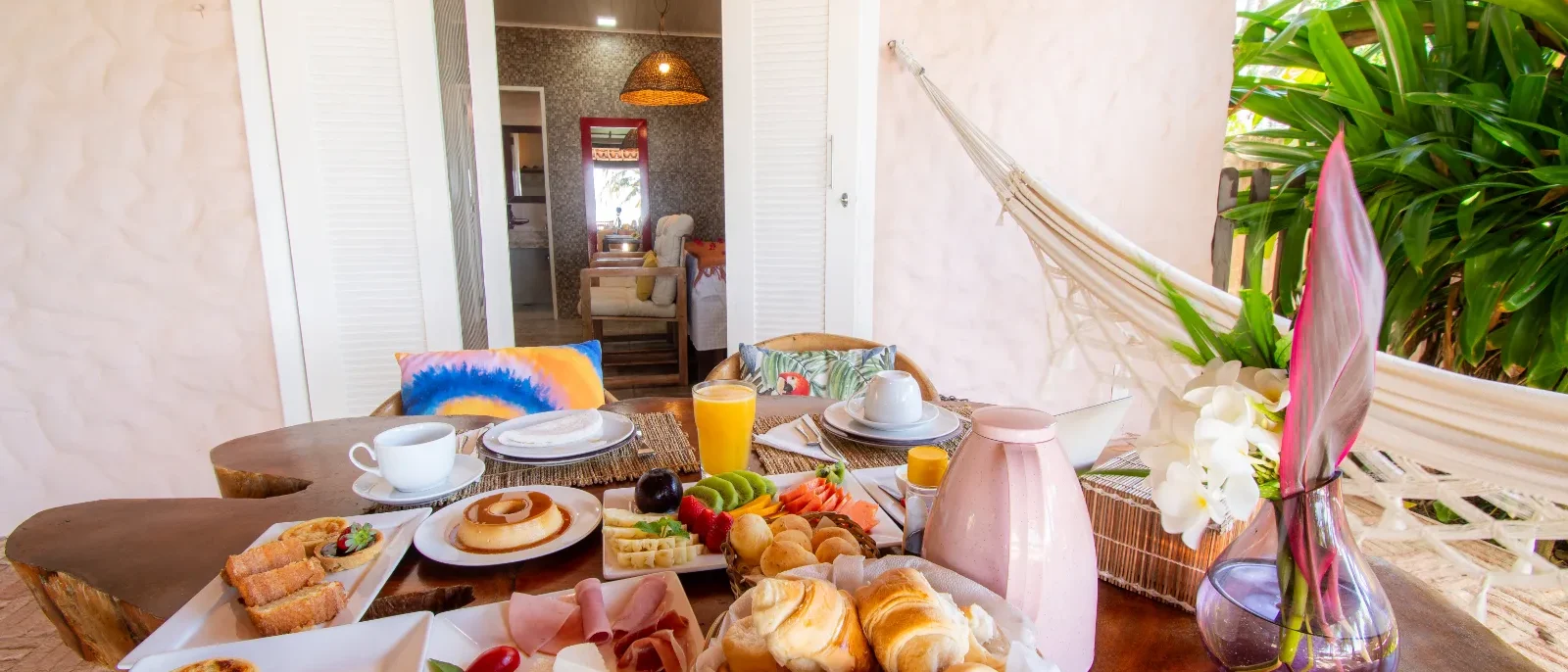 Mesa com café da manhã contendo pães, croissants, frutas, presunto, sucos, café e flores, com porta aberta ao fundo mostrando interior da casa