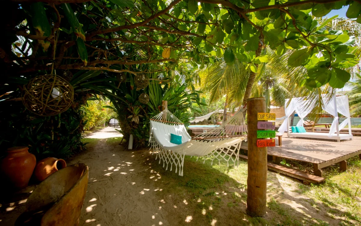 Área sombreada por vegetação tropical com redes brancas penduradas, plataformas de madeira, cabanas brancas ao fundo e decorações coloridas em postes de madeira