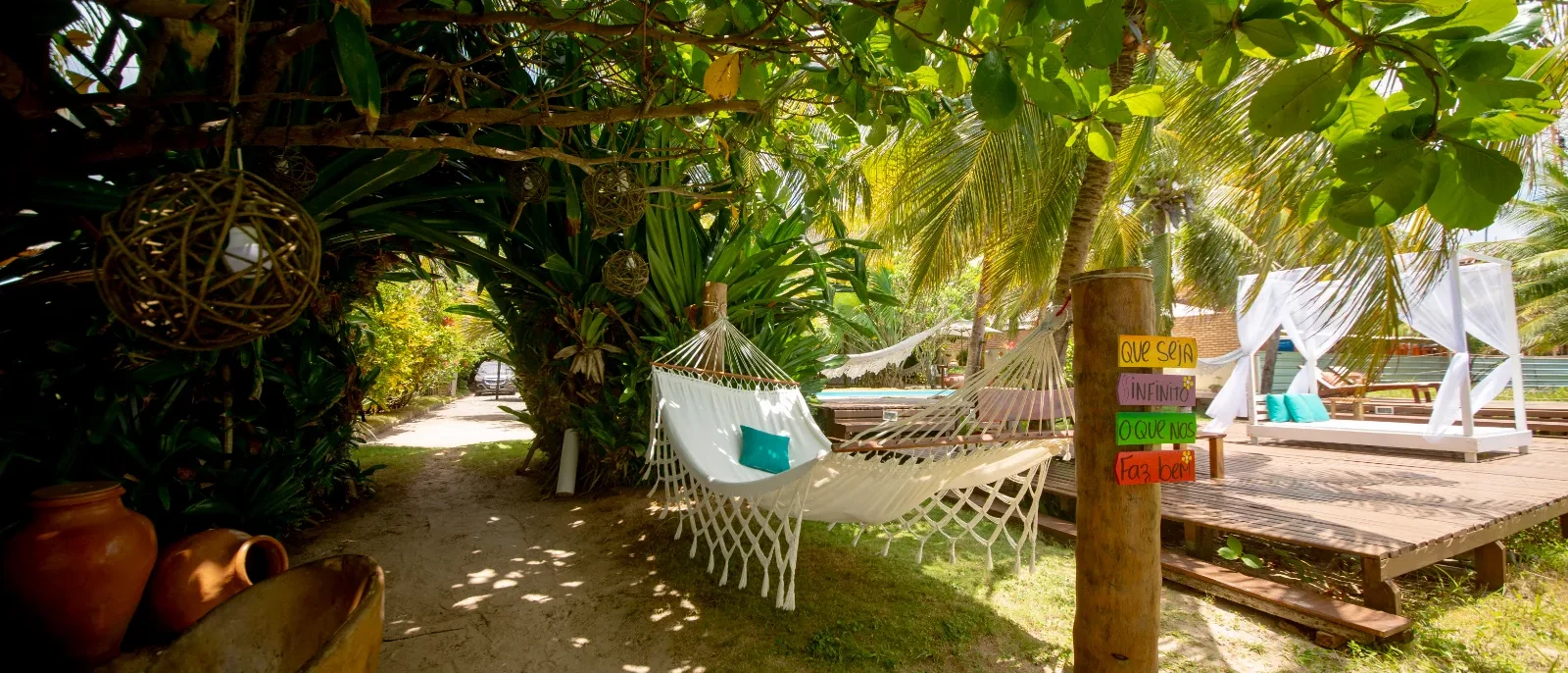 Área sombreada por vegetação tropical com redes brancas penduradas, plataformas de madeira, cabanas brancas ao fundo e decorações coloridas em postes de madeira