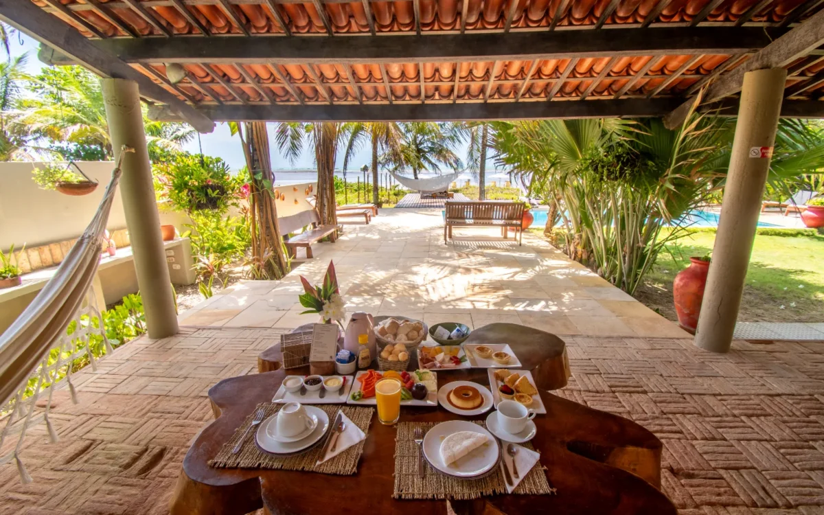Mesa com café da manhã servido sob pérgolado com telhas de terracota, vista para jardim com palmeiras, piscina e estrutura de lazer ao fundo